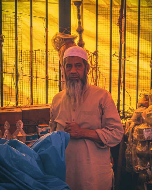 Interior view of a home or marketplace setting with a yellow-tinted plastic or fabric sheet ceiling, supported by metal poles. In the foreground, a man dressed in a light-colored traditional shirt and