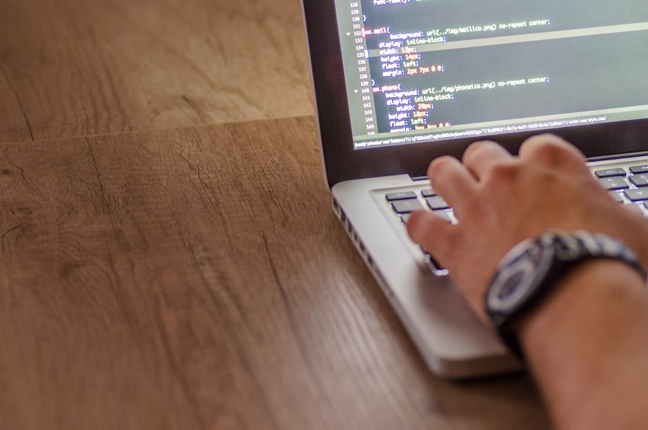 A person's hand, wearing a patterned wristwatch, is typing on a silver laptop placed on a wooden surface. The laptop screen displays a code editor with multiple lines of HTML or similar programming la