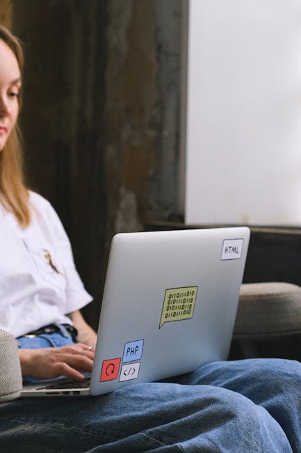 A person's hand, wearing a patterned wristwatch, is typing on a silver laptop placed on a wooden surface. The laptop screen displays a code editor with multiple lines of HTML or similar programming la
